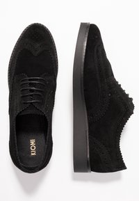 Brogues neri in suede con lacci presentano intricate perforazioni, una superficie testurizzata e una suola in gomma piatta con un leggero rialzo. Branding dorato "KIOMI" all'interno.