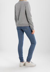 Persona vestida con jeans ajustados azules, suéter gris de manga larga y zapatillas blancas, de pie con la espalda hacia la cámara.