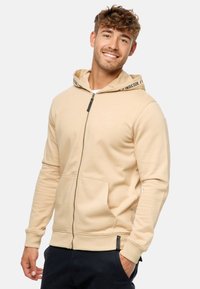 Beige zip-up hoodie i blødt stof med frontlommer, justerbar snørehætte og et logo på brystet; har kontrasttekst på hætten.