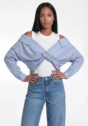 Kobieta o długich czarnych włosach nosi biały top bez rękawów, niebieską koszulę w paski zawiązaną poza ramionami oraz niebieskie dżinsy z wysokim stanem, ręce na biodrach.