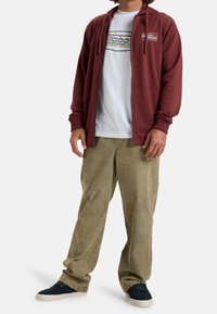 Sudadera con cremallera color marrón encima de una camiseta blanca con logo, combinada con pantalones de pana en un suave tono verde oliva y zapatillas de deporte azul marino.