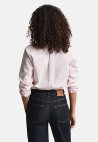 Cămașă roz cu dungi și mâneci suflecate, asortată cu blugi albastru închis, având o etichetă de piele cu logo și cusături contrastante pe buzunare.