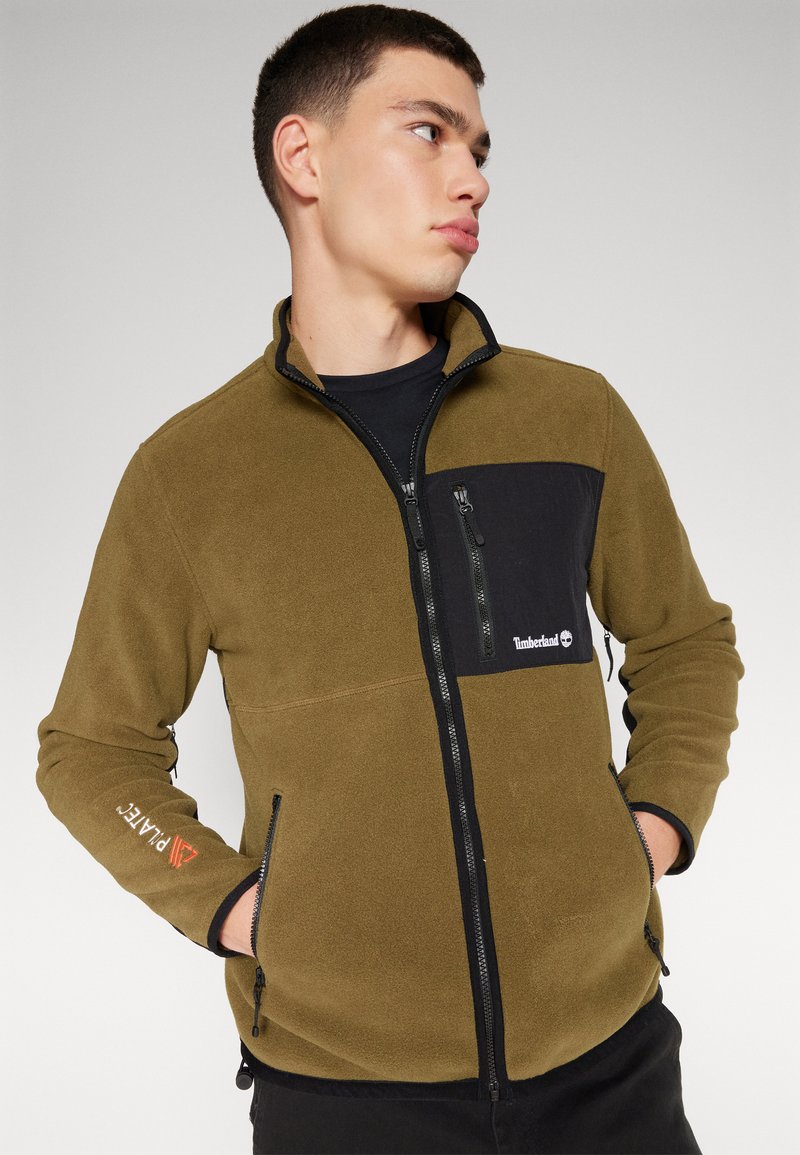 Oliwkowa polarowa kurtka z czarną kieszenią na piersi, zamkami błyskawicznymi i akcentami logo. Posiada wysoki kołnierz i boczne kieszenie do przechowywania.