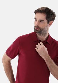 Burgunderfarbendes Poloshirt mit flachem Kragen, kurzen Ärmeln und einem Knopf am Hals. Der Stoff wirkt glatt und leicht, mit einer figurschmeichelnden Passform.