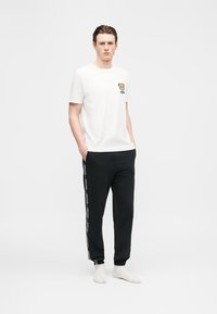 Homme portant un t-shirt blanc avec un petit logo d'ourson et un pantalon de jogging noir avec une bande logo blanche, debout les mains dans les poches.
