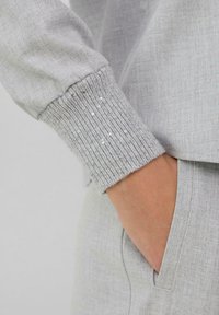 Hand in der Tasche mit einem grauen Strickpullover mit langen Ärmeln, der einen gerippten Bund hat, der mit kleinen weißen Pailletten verziert ist.