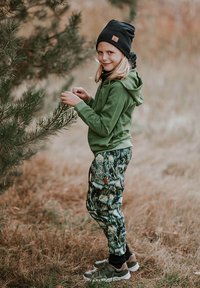 Zielona bluza z kapturem, wzorzyste spodnie dresowe z motywem lasu, czarna czapka typu beanie i tenisówki. Otoczenie na zewnątrz z trawą i drzewami.