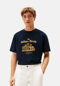 Joven vestido con una camiseta azul marino de Tommy Hilfiger con un horizonte urbano dorado y pantalones blancos, de pie frente a un fondo neutro.