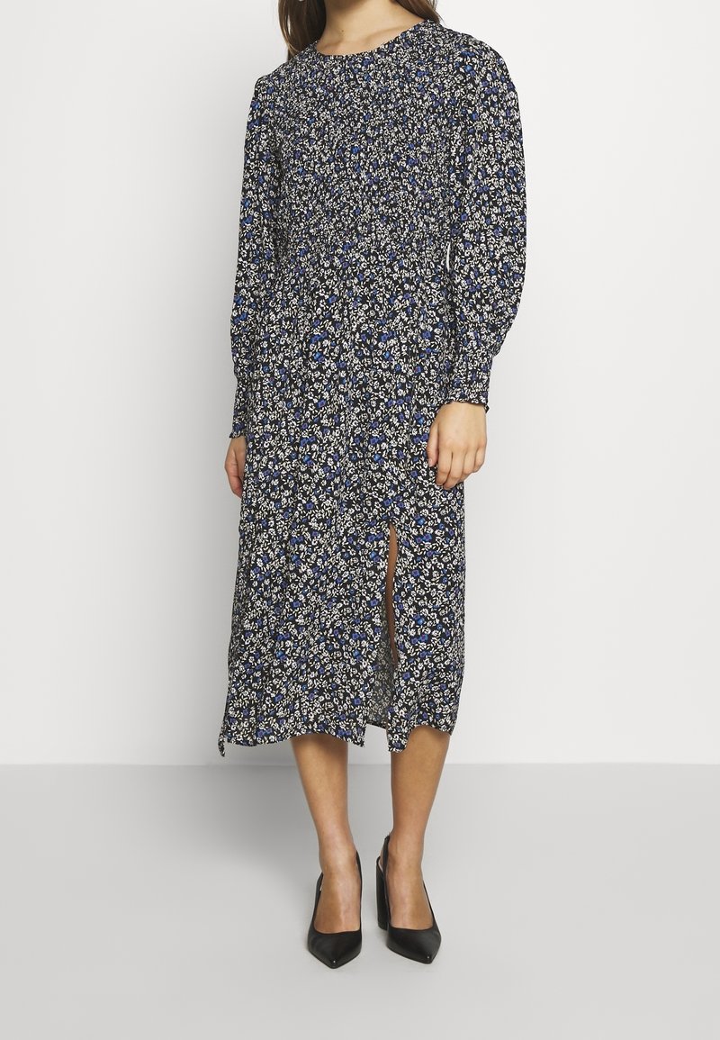 Pitkä kukkamekko, jossa pitkät hihat, tummansininen tausta, pienet valkoiset ja siniset kukat sekä reidestä korkea halkio. Yhdistetty mustiin teräväkärkisiin korkokenkiin.