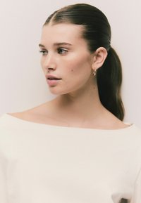 Porträt einer Person mit glatter Haut, die ein weißes schulterfreies Oberteil und goldene Creolen trägt, mit glattem, dunklem Haar.