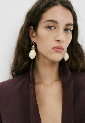 Femme aux longs cheveux foncés portant de grandes boucles d'oreilles crème texturées et un haut prune foncé à col haut, regardant légèrement de côté avec une expression neutre.