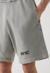 Pantalones cortos de algodón gris con una cinturilla elástica, bolsillos laterales y un logo negro en la pierna izquierda, que muestran una textura suave y un diseño casual.