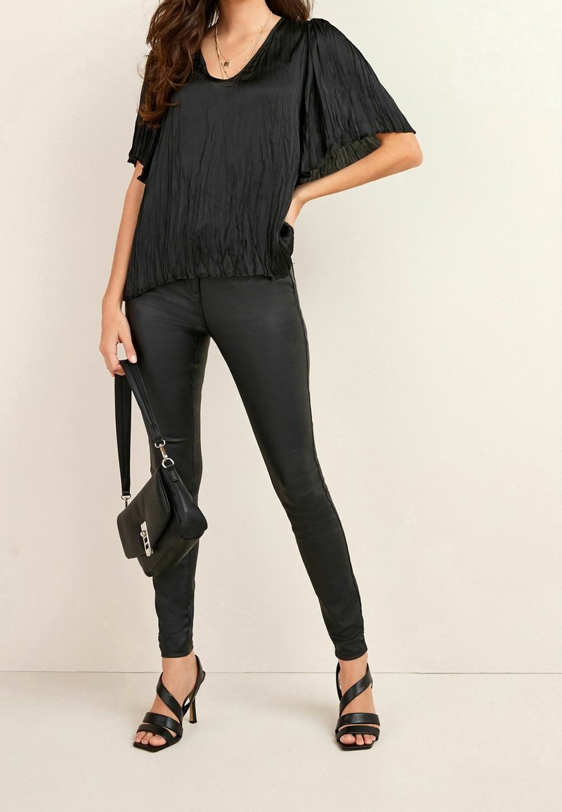 Femme portant un haut court en tissu froissé noir à manches courtes, des leggings en cuir noirs, des sandales à talons hauts noires et tenant un sac à main noir.