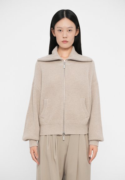 Beige strikket lynlås sweater med høj krave, to forlommer og store ærmer, lavet af et tekstureret garnmateriale.