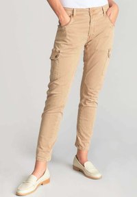 Pantalones cargo beige con corte ajustado, confeccionados en suave algodón, que cuentan con dos bolsillos laterales, cierre frontal con botón y detalles de costura visibles.