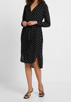 Sort langærmet polkaprik kjole med V-hals og frontslids, båret med sorte remsandaletter med lav hæl.