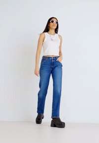 Top sin mangas blanco, jeans rectos azul medio y zuecos de plataforma negros. Los accesorios incluyen unas gafas de sol y un collar de mariposa rosa.