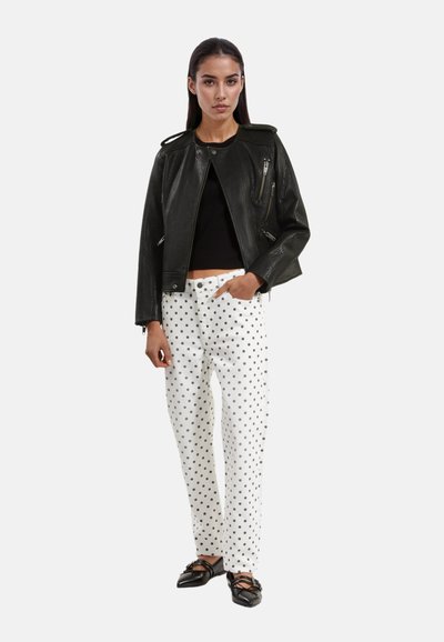 Zwarte leren cropped jas met schouderepauletten, gecombineerd met witte broek met stippen en zwarte schoenen. Gladde textuur en een getailleerd ontwerp.