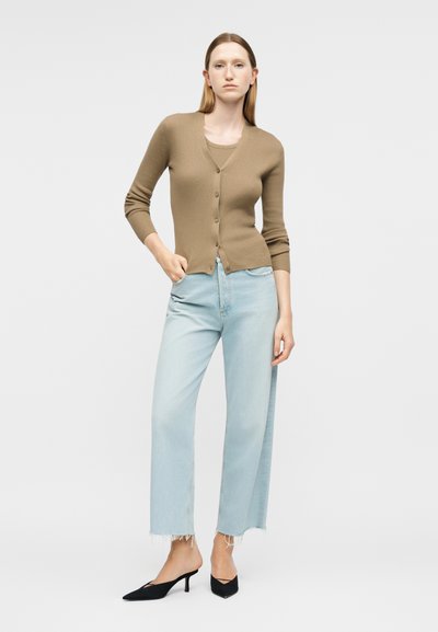 Γυναίκα που φοράει ένα καφέ ζακέτα με κουμπιά, ανοιχτό μπλε κοντά τζιν με ξεφτισμένα τελειώματα και μαύρες γόβες με λουράκι πίσω και μυτερό μύτη, στέκεται μπροστά σε ένα απλό φόντο.