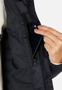 Giacca puffer nera con una tasca con zip, che mostra uno smartphone parzialmente inserito. Il tessuto ha una consistenza liscia e un leggero lucore.