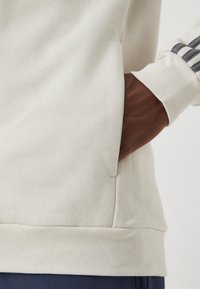 Pulôver bege claro com um acabamento texturizado, apresentando um bolso lateral e punhos canelados. Vem com detalhes em azul marinho nas mangas.