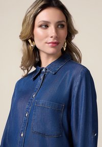 Blue denim shirt with a collared neckline, front buttons, and a chest pocket. Soft texture with subtle sheen. Gold statement earrings.