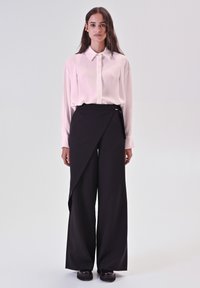 Camicia bianca con maniche lunghe abbinata a pantaloni neri a vita alta e gamba larga con un design avvolgente asimmetrico e un accento laterale.