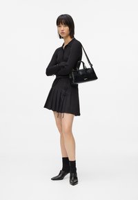 Γυναίκα που στέκει με τα χέρια σταυρωμένα, φορώντας έναν μαύρο ριγέ πουλόβερ, πλισέ φούστα με δαντελένιες λεπτομέρειες, μαύρες κάλτσες, παπούτσια με τακούνι και κρατώντας μια μαύρη τσάντα.