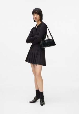 Donna in piedi con le braccia incrociate, indossa un maglione a coste nero, una gonna plissettata con dettagli in pizzo, calze nere, scarpe col tacco e porta una borsa nera.