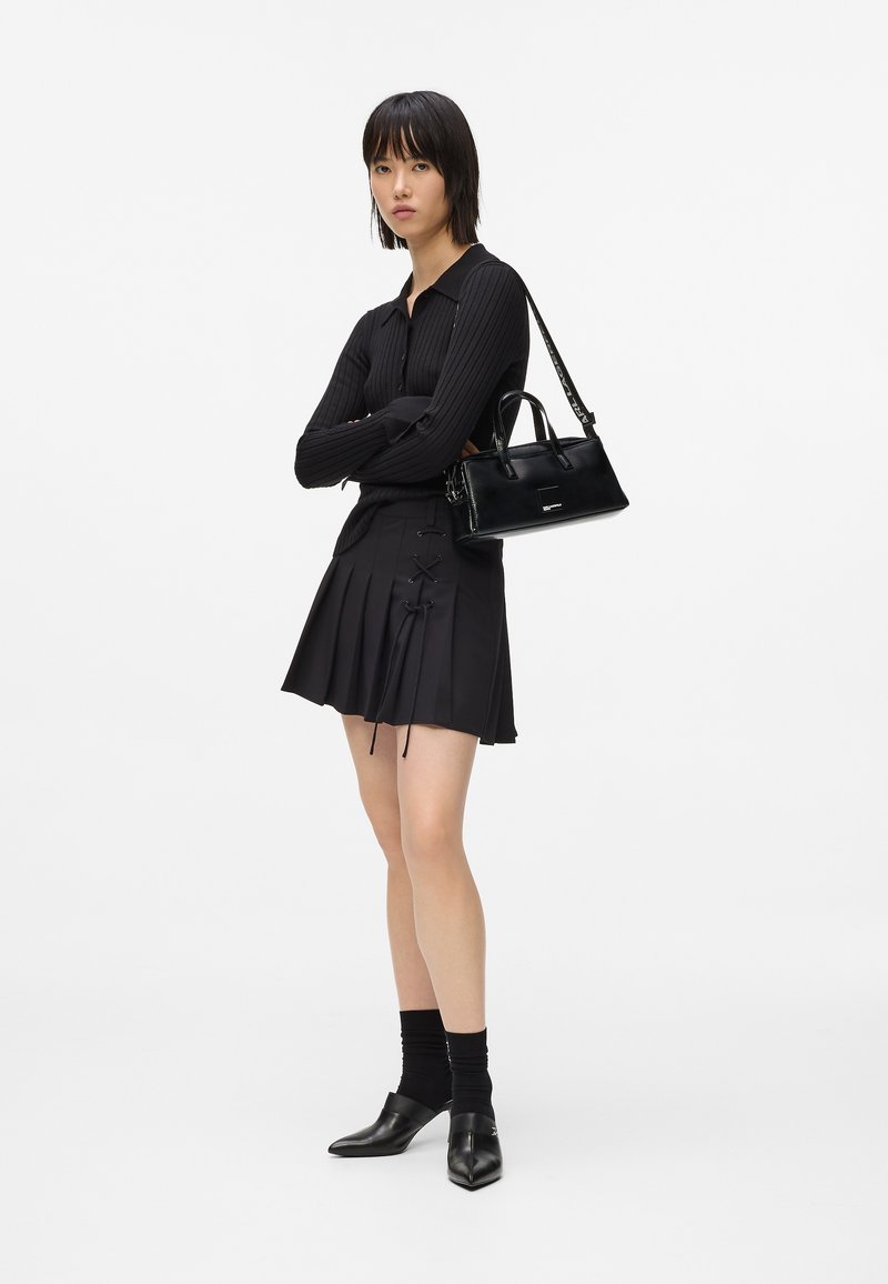 Γυναίκα που στέκει με τα χέρια σταυρωμένα, φορώντας έναν μαύρο ριγέ πουλόβερ, πλισέ φούστα με δαντελένιες λεπτομέρειες, μαύρες κάλτσες, παπούτσια με τακούνι και κρατώντας μια μαύρη τσάντα.