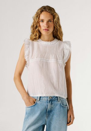 Frau mit welligem blondem Haar, die eine ärmellose weiße Bluse mit gerafften Schultern und hellblaue, hoch taillierte Jeans trägt, stehend mit einer Hand in der Tasche.