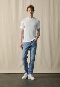 Hvit t-skjorte med korte ermer, lyseblå jeans, hvite sneakers med teksturert såle, stående mot en nøytral beige bakgrunn.