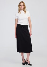 Top ajustado de manga corta blanca, falda larga negra con detalle de cinturón y zapatos de tacón bajo negros con puntera cuadrada. Texturas lisas en todo.