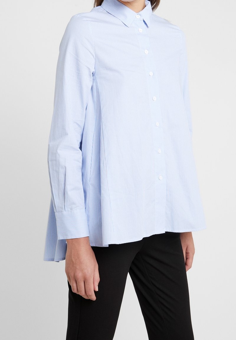 Woman wearing a light blue long-sleeve button-up shirt with thin white stripes and black pants, standing against a plain background.