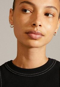 Primer plano de un rostro que lleva un suéter negro de canalé con costuras blancas. Presenta pequeños pendientes de aro en plata y una textura de piel suave y natural.