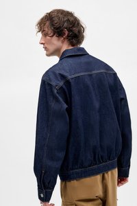 Donkerblauwe denimjas met een ontspannen pasvorm, voorzien van een kraag, elastische taille en knoopmanschetten. Glad textuur met zichtbare naden.