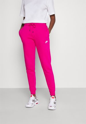 Persoon gekleed in felroze Nike joggingbroek met trekkoord, witte sneakers en een effen wit T-shirt met korte mouwen, staand tegen een neutrale achtergrond.