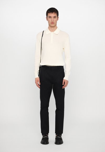 Camisa polo de manga longa, cor creme, com colarinho, combinada com calças pretas ajustadas e mocassins pretos. Design minimalista, texturas suaves.