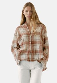Langärmliges, knöpfbares Hemd in einem karierter Muster aus Beige, Rot und Orange, mit entspannter Passform und Kragen. Getragen mit weißen Hosen.