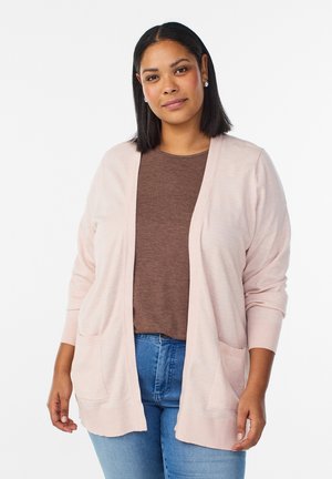 Frau mit schulterlangem dunklem Haar trägt hellrosa offenen Cardigan, braunes Top und blaue Jeans vor weißem Hintergrund.