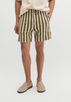 Mann trägt beige und olivgrüne Shorts mit vertikalen Streifen und Kordelzug, beige Hemd und beige Slip-on-Schuhe, steht mit den Händen in den Taschen.