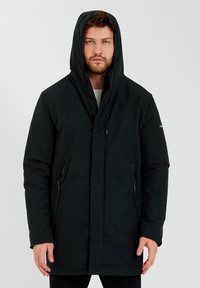 Zwarte, lange pufferjas met capuchon, voorzien van ritszakken en een zachte textuur. Minimalistisch ontwerp zonder zichtbare patronen of logo's.