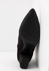 Botte en daim noir avec bout pointu et semelle texturée, positionnée debout contre un mur d'angle blanc.