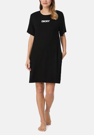 Woman wearing a black knee-length DKNY dress with short sleeves, standing barefoot against a plain white background.