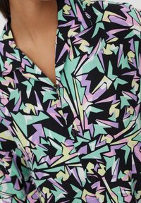 Chemise à col avec boutons et motif géométrique coloré en vert pastel, violet, jaune et blanc sur fond noir.
