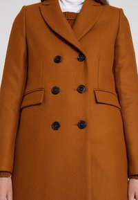 Double-breasted brown coat made of textured fabric, featuring black buttons, notched lapels, and pockets on both sides.