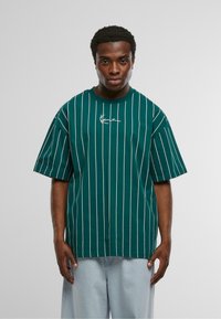 Zaļš svītrainais T-krekls ar brīvu piegriezumu, kam ir baltas vertikālas līnijas un roku rakstīts logo uz krūtīm. Mīksts kokvilnas materiāls.