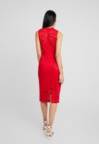 Robe sans manches en dentelle rouge avec une silhouette ajustée, longueur genou, comportant une fente au dos et une taille marquée. Portée avec des talons blancs ouverts à l'avant.