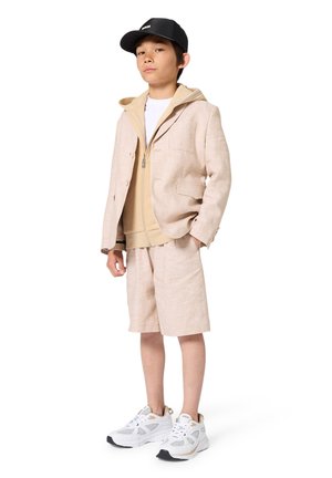 Beige Leinen-Sakko über einem beigen Hoodie, kombiniert mit passenden Shorts. Weiße Sneakers mit grauen und goldenen Akzenten und eine schwarze Kappe.