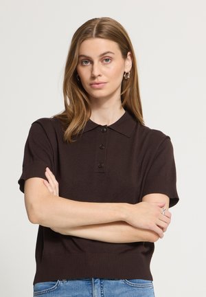 Femme aux cheveux châtain clair portant un polo à manches courtes marron foncé et un jean bleu, bras croisés, expression neutre, fond clair uni.